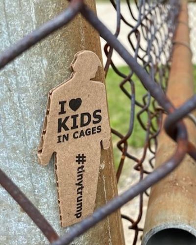 a photograph of a tiny trump with the slogan 'I love kids in cages' shown stuck to the post of a chain link fence