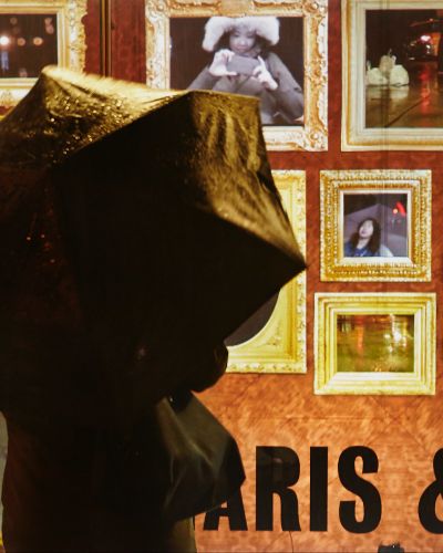 Photograph of someone with an umbrella, outside in the rain, looking at a gallery storefront which shows a projection of nearly a dozen golden-framed video feeds, some of which show different facets of the viewer (her face, shoes, torso), some of which show facets of the urban landscape in the background (sidewalk, street, cars, etc.).