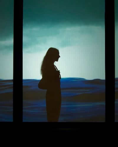 Nighttime photograph showing a urban storefront showing a video of a contiguous body of water projected across 4 of its windows; lower half of the project is dark blue water, the upper half is light blue sky