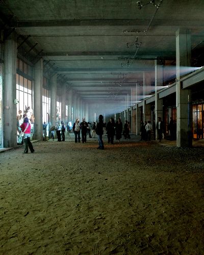 Photograph of a relatively dark, cavernous space (like the inside of a large, empty commercial building) with art festival goers milling about, some of whom have climbed into the windows to interact with what's being projected onto them (the water scene described above). On the upper left of the photograph one can see a line of about a dozen projectors shining light across the empty space onto the windows.