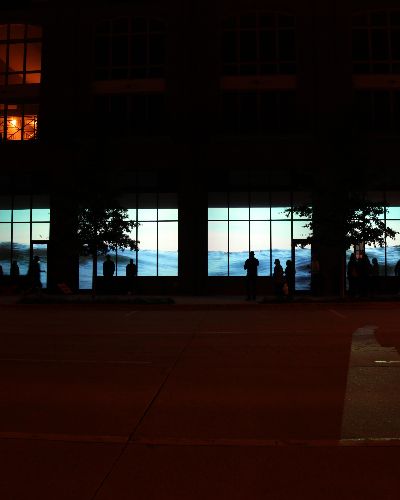 Nighttime photograph of a large commercial center with six of its street-level storefront windows showing a contiguous video projection of water ebbing and flowing.
