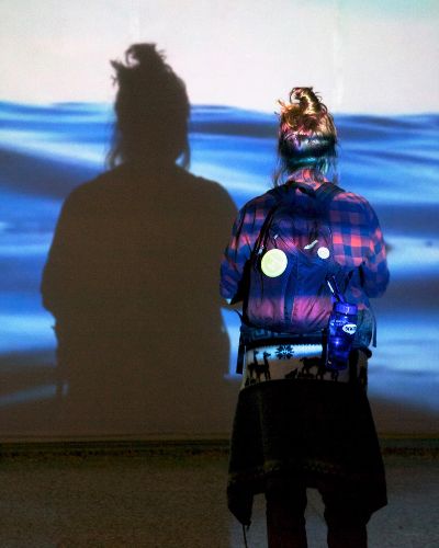 Photograph of the back of a seemingly teenage girl wearing a backpack staring at the life-size projection of water in front of her. We see her shadow in the projection of the water.
