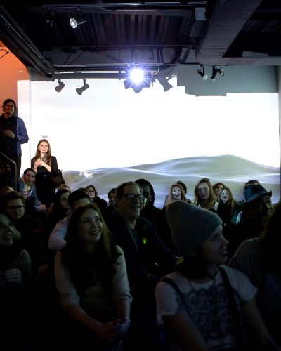 Photograph of a crowd of people sitting down in a dark gallery space with a body of water projected on the walls behind
