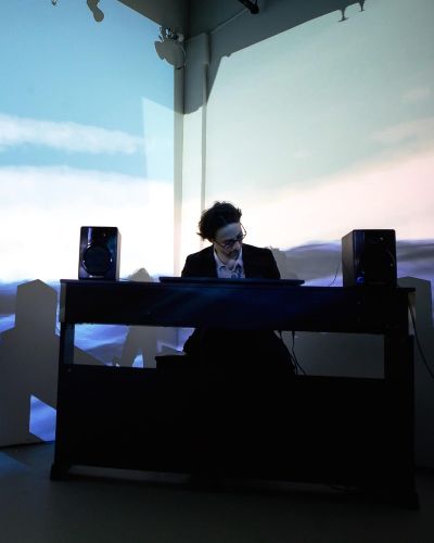Photograph of the artist, Eric Corriel, swaying while playing piano in the corner of a gallery, which is mostly dark. Projected onto the two walls that form the corner is his piece, Water Will Be Here, which I'm sure you've guessed by now, consists of a video of a body of water.