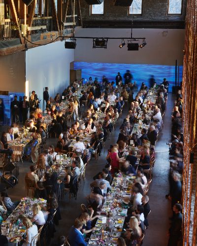 Overhead shot of a gala event&mdash;dozens of elegantly decorated tables with white table cloths and well-dressed people sitting at them. Behind them, on a long white wall, a video of a body of water is projected