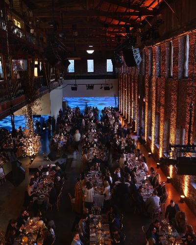 Overhead shot of a gala event, over elegantly decorated tables with well-dressed people seated at them. Behind them, on a long white wall, a video of a body of water is projected 