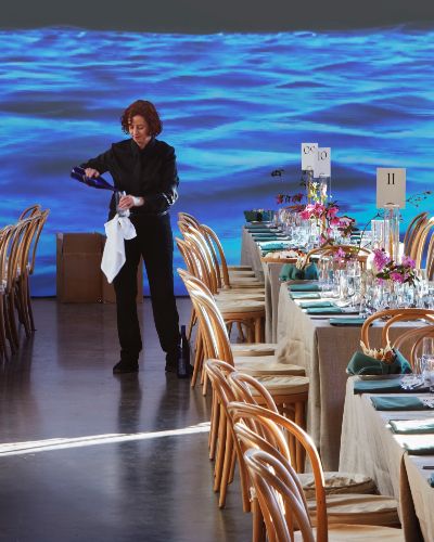 Foreground: white table clothed tables with numbers suggesting a gala event, though no one is seated at them yet. Middleground: a server pours water into a glass. Background: a video of a body of water is projected on a white wall.