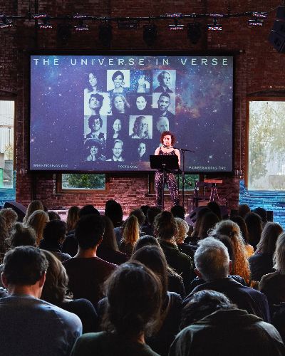Photograph of an event in which approximately a hundred people are seated&mdash;we see the backs of their heads. In the background is a woman on stage, reading (a poem). Behind her is a screen with text that reads 'The Universe In Verse,' which is flanked on both sides by a projection of video of water on a brick wall.