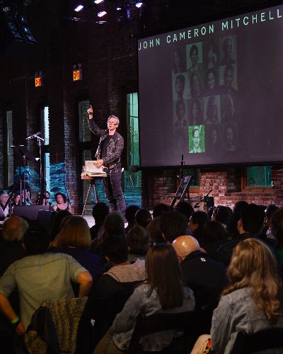 John Cameron Mitchell gestures with a finger raised to the sky as he reads a poem to an audience. A video of a body of water is projected onto the interior brick wall behind him.