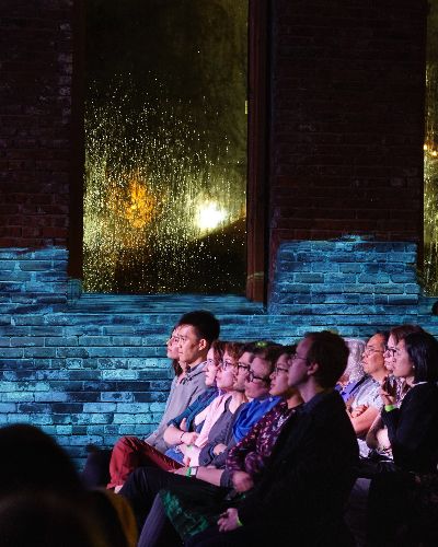 Photograph of three rows of audience shot in profile. A video of a body of water is projected on the brick wall behind them.