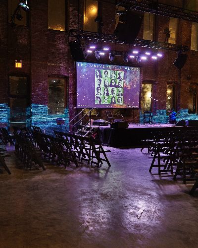 Photograph of a large empty room with over a hundred empty folding chairs. A small stage is in the center and above it, a screen indicates the name of the event: 'The Universe In Verse.' A video of a body of water is projected on the brick wall in the background.