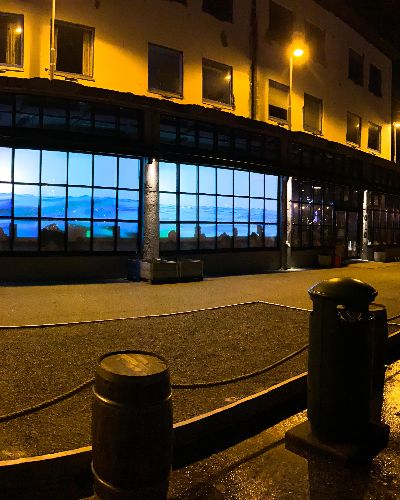 Panoramic photograph with Water Will Be Here - a video installation projected into the windows of a bar showing water ebbing and flowing slowly - on the left and several spectators outside of the bar on the right, with a body of water behind them