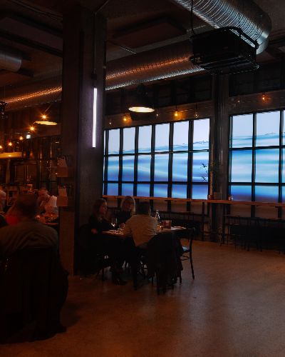 Interior photograph showing people at a bar doing normal bar stuff with Water Will Be Here projected in the windows behind them