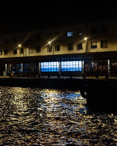 Wide shot showing Water Will Be Here (same installation as above) projected into windows of a bar about 150 feet away on the right. On the left is a body of water that is very close to the bar.