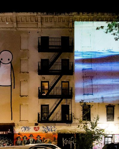 Photograph of a white building in New York City which has a 35-foot-tall graffiti drawing of a stick figure reaching towards the sky on the left and Water Will Be Here projected, equally as tall, on the right