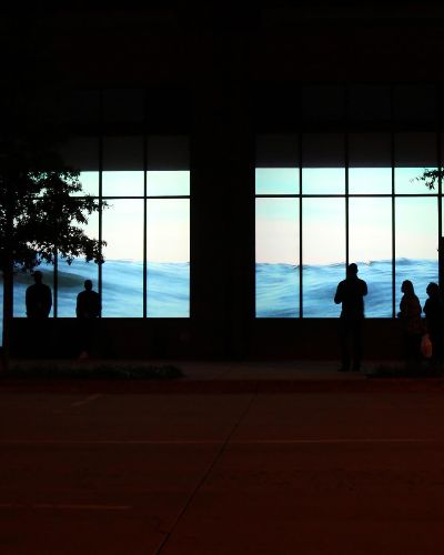 Nightime photograph of a several storefronts with a projection of water (from the sea) in their windows. Water level is contiguous across many storefronts. Inside the storefronts are silhouettes of people interacting with the video installation.