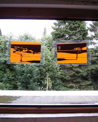 Photograph of two artworks, encased in transparent plexiglass, hanging in a large open window. Each artwork contains an element of a diptych, which taken together form a golden/yellow-ish abstract landscape with one tree on each side. Several trees are visible behind the hanging artworks in the open window.