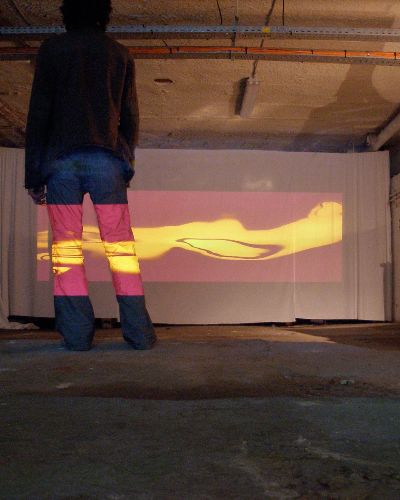 Photograph of the back of a person, wearing blue jeans and a dark jacket, with a projection of abstract burgundy and yellow organically shaped forms that are also projected onto a large screen at the end of an industrial-looking space.