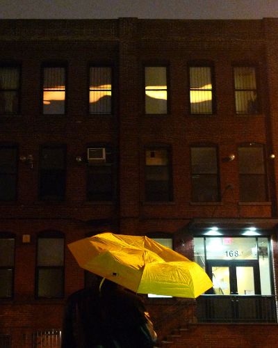 Nighttime photograph of a large, three-story industrial brick building. In the top floor, four center windows display a projection of an abstract, fluid yellow band. In the foreground is a person standing with a yellow umbrella.