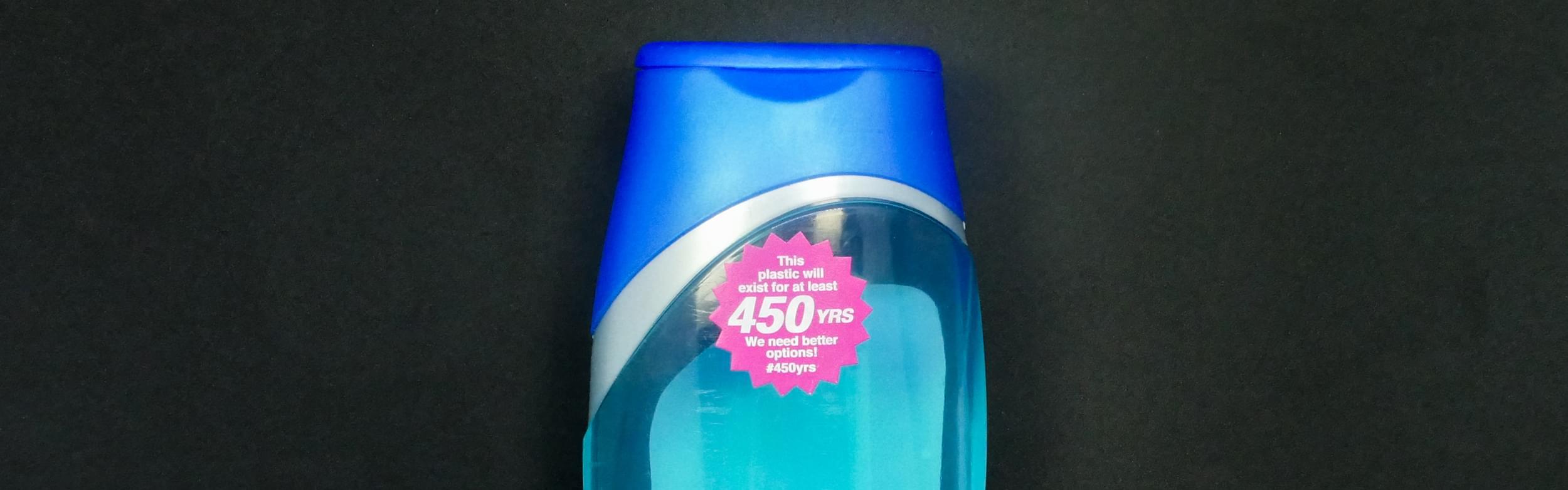 Close-up photograph of a blue plastic bottle, shaped like a liquid soap container. On the front of the bottle is a magenta, sunburst-shaped sticker that says, 'This plastic will exist for at least 450 YRS. We need better options! #450yrs.'