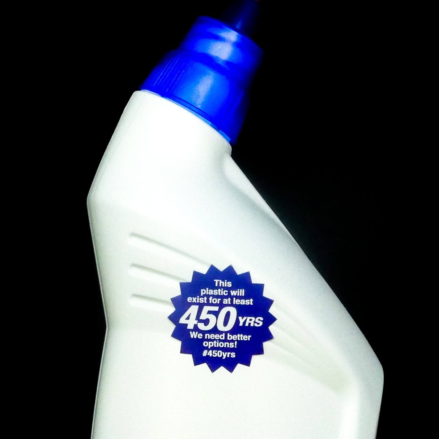 Close-up photograph of a white plastic bottle, shaped like a toilet cleaner container. On the front of the bottle is a blue, sunburst-shaped sticker that says, 'This plastic will exist for at least 450 YRS. We need better options! #450yrs.'