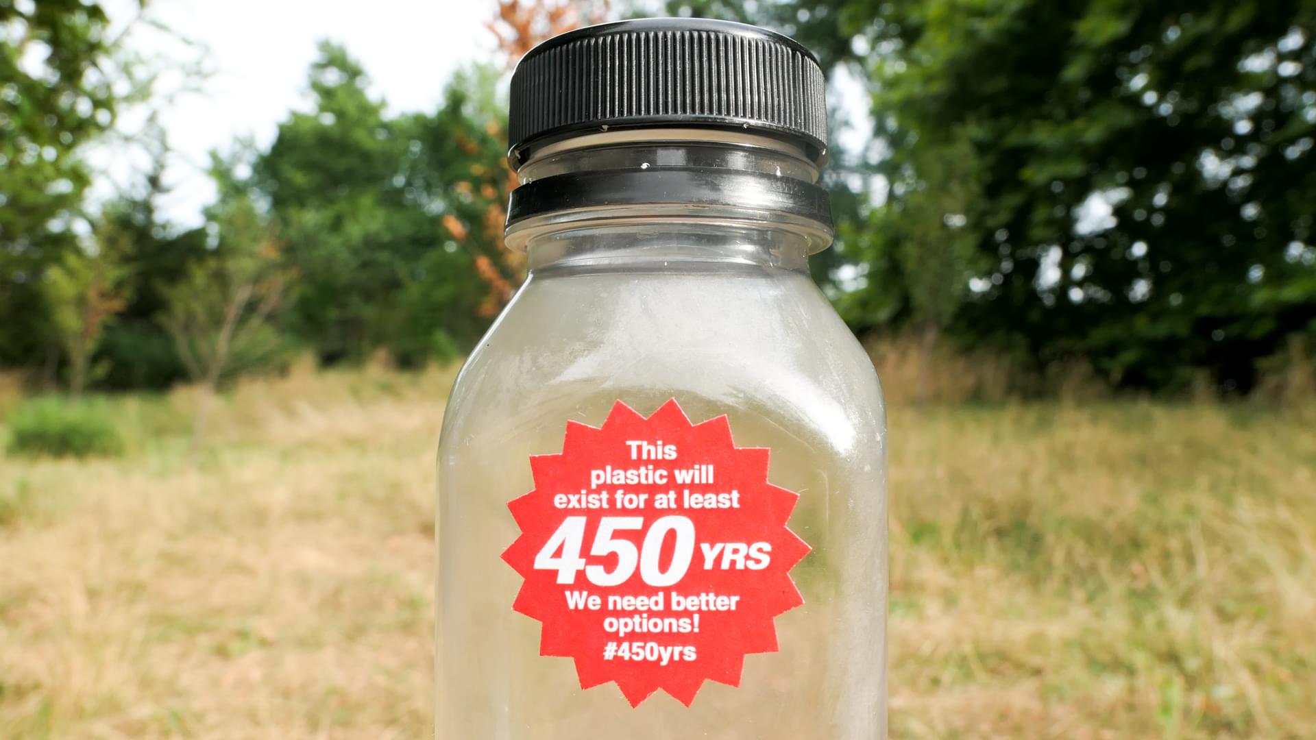 A plastic bottle with red, starburst-shaped sticker on its front that reads: 'This plastic will exist for at least 450 YRS. We need better options! #450yrs. In the background is a blurry green field.