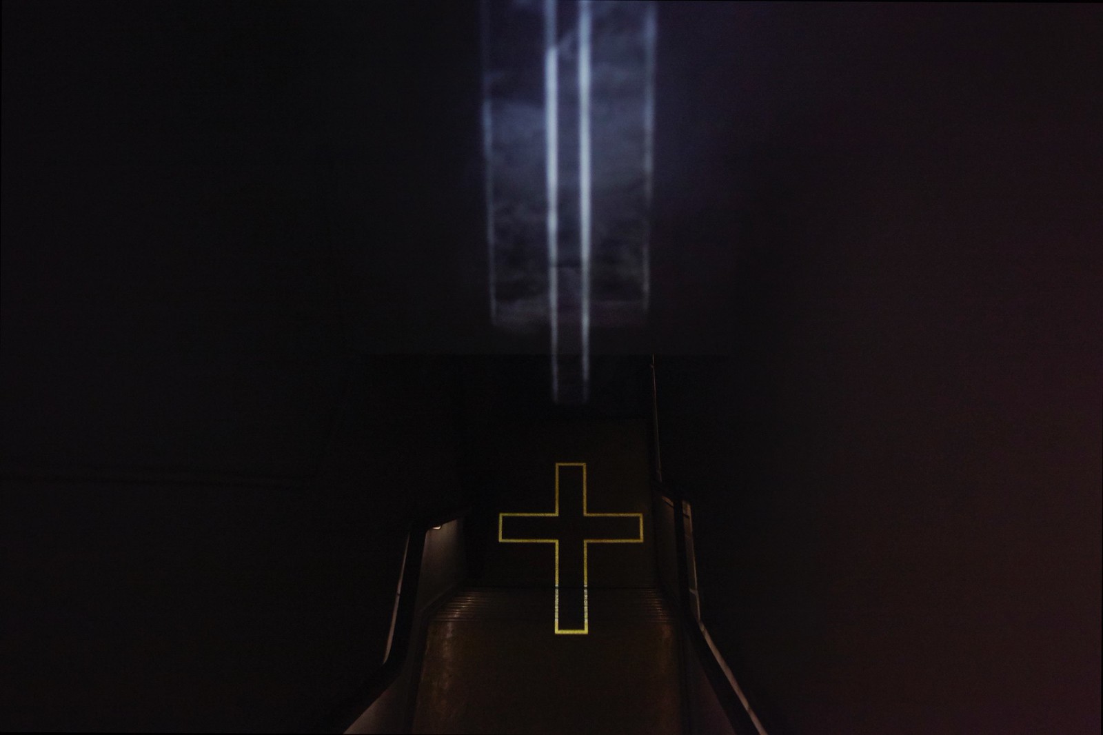 Photograph taken from atop a dark stairwell showing the outline of a crucifix projected onto the floor at the bottom of the stairwell along with the vertically oriented beams of light, visible in midair, that constitute it.