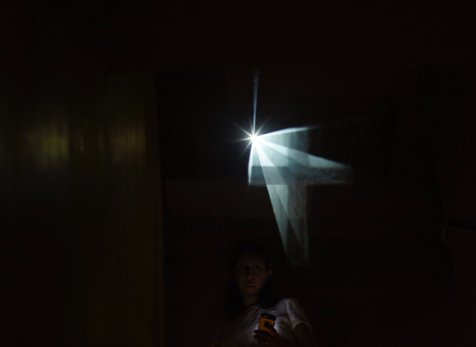 Photograph of a barely lit room with a person taking a selfie at the bottom-middle of the frame and a clearly defined crucifix seemingly floating above her left shoulder. The floating crucifix emanates from a singular point of bright light in the middle of the frame.