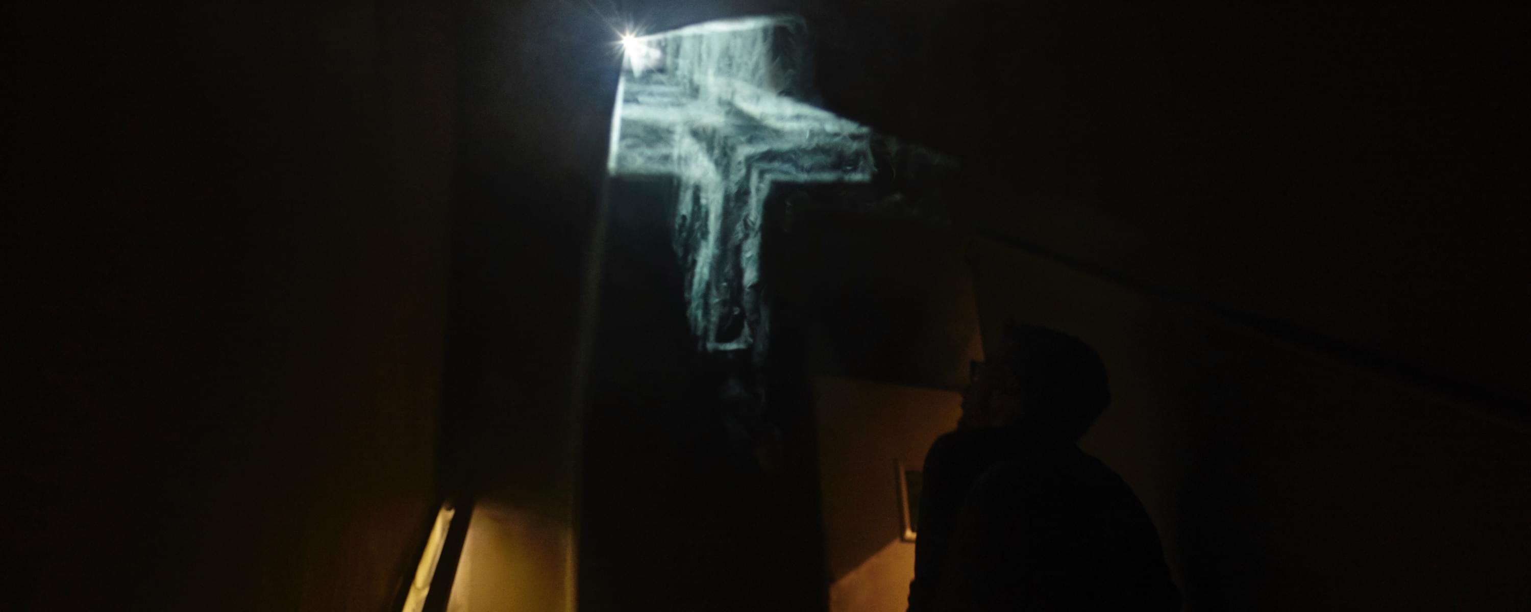 Photograph; it is extremely dark, on the right is an inkling of an illumined handrail indicating a staircase. In the middle is a floating, cloudy, illuminated crucifix in three dimensions; towards the bottom right, if one squints, one can make out a viewer in the darkness looking up at the ghostly crucifix