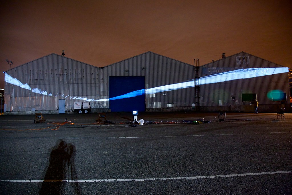 Nighttime photograph showing a large warehouse in the background onto which a large oval of white rectilinear geometric forms is projected. In the foreground is a large, empty parking lot with projection equipment strewn about.