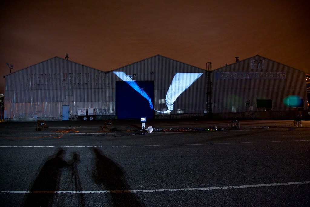 Nighttime photograph showing three large warehouses in the background onto which is projected a few dozen white butterfly-like forms, one behind the other in a figure-eight-ish configuration. The figure-eight-ish configuration is not flat but rather rotated along the y-axis in 3D space. In the foreground is a large, empty parking lot with projection equipment strewn about, and the sky behind the warehouses is a dark shade of burnt orange.