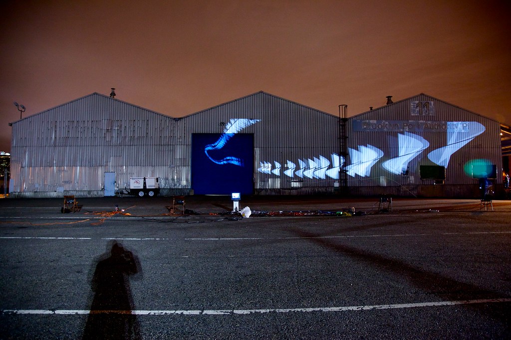 Nighttime photograph showing three large warehouses in the background onto which is projected over a hundred distinct white geometric forms, one in back of the other in a very long, slightly arched line. In the foreground is a large, empty parking lot with projection equipment strewn about, and the sky behind the warehouses is a dark shade of burnt orange.