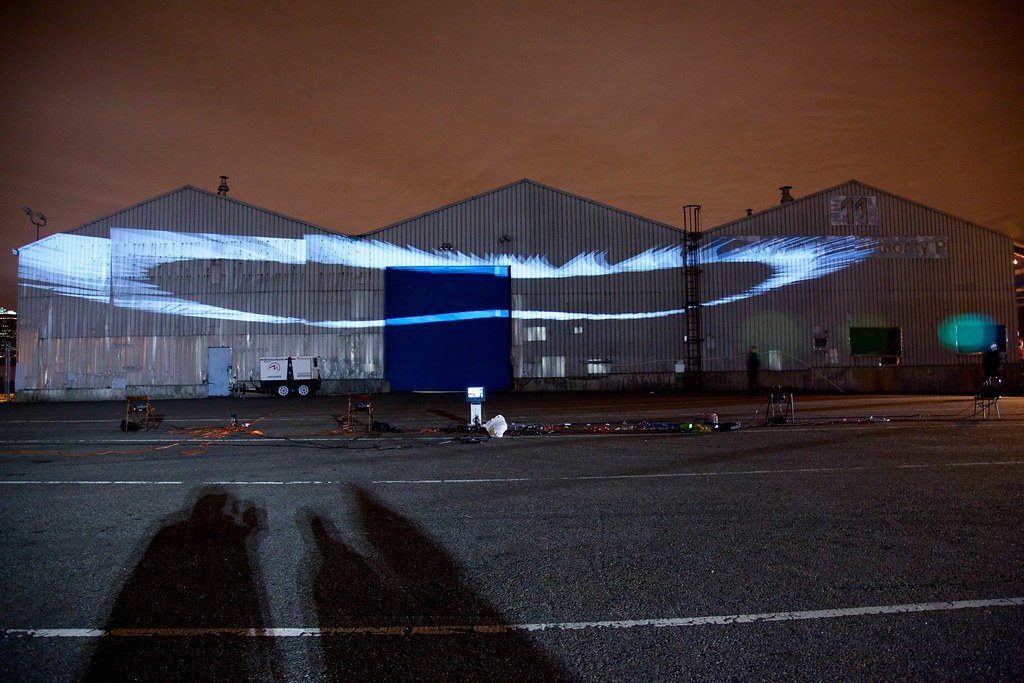 Nighttime photograph showing three large warehouses in the background onto which is projected several dozen white oval-esque forms, one behind the other in a flying-V formation. The flying-V formation is not flat but rather angled downward. In the foreground is a large, empty parking lot with projection equipment strewn about, and the sky behind the warehouses is a dark shade of burnt orange