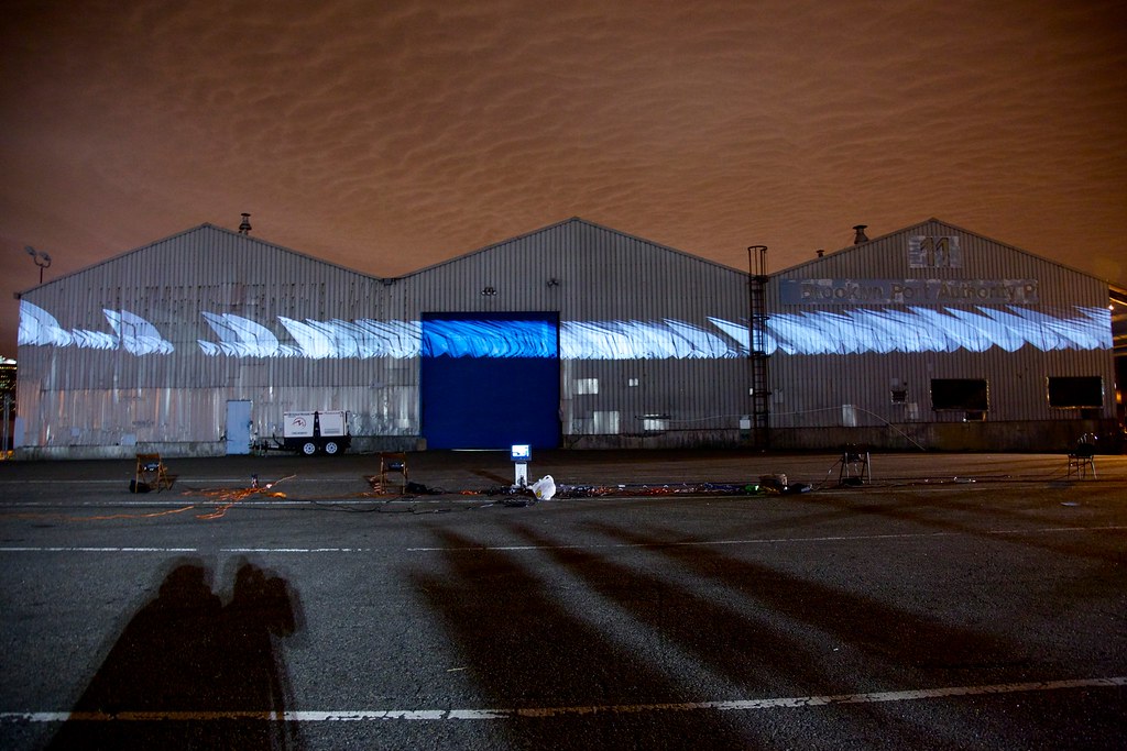 Nighttime photograph showing three large warehouses in the background onto which is projected a large white, swoosh-like form. In the foreground is a large, empty parking lot with projection equipment strewn about, and the sky behind the warehouses is a dark shade of burnt orange.