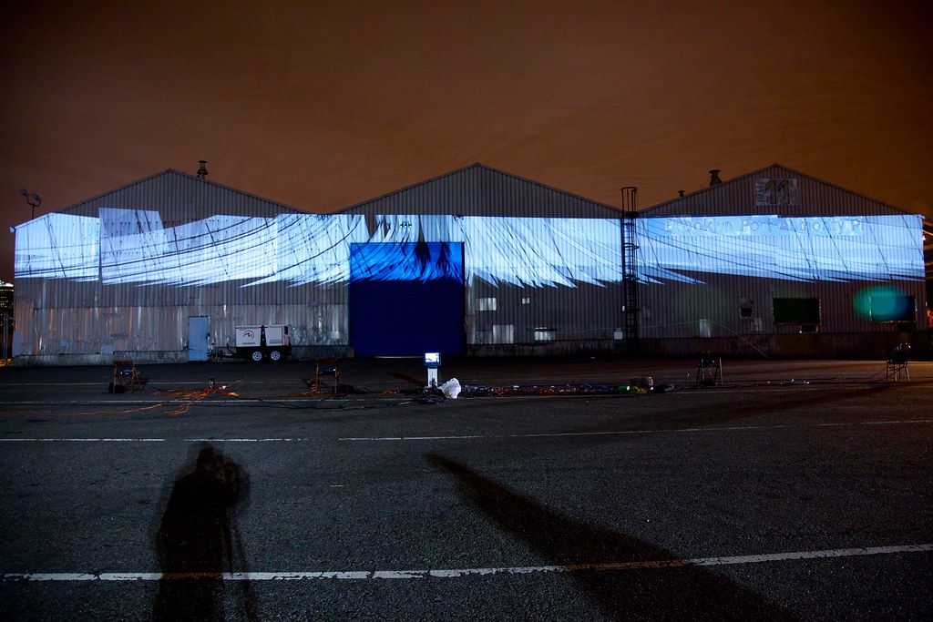 Nighttime photograph showing three large warehouses in the background onto which is projected over 100 white sail-shaped forms, one behind the other along a cinched oval-shaped path. In the foreground is a large, empty parking lot with projection equipment strewn about, and the sky behind the warehouses is a dark shade of burnt orange.