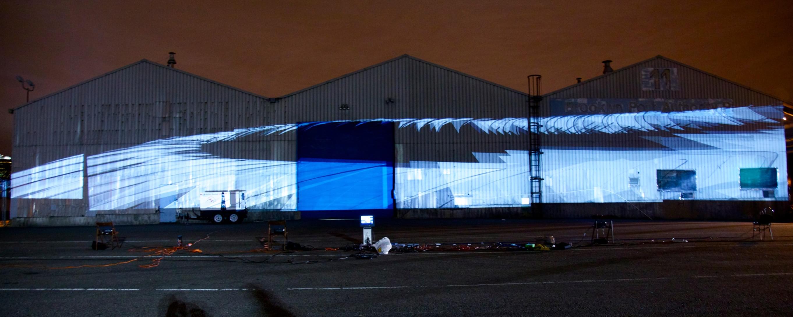 Nighttime photograph of three large warehouses, each with a triangular house-like top, with a large, 200+ foot projection of what looks like a series of graphically designed white sails following each other around a circular loop.