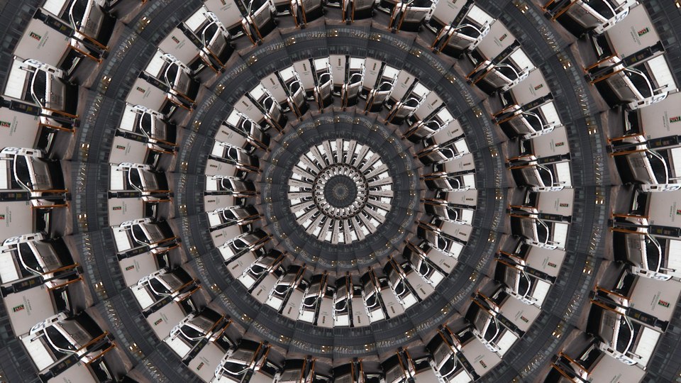 Algorithmically generated kaleidoscopic image consisting of a series of infinite-mirror-esque concentric discs, each one subdivided into multiple tranches that show the same image, albeit rotated by a consistent amount across the disc. In this case, the image that gets repeated across all discs at different angles is a photograph of a white SUV in front of a white delivery truck.