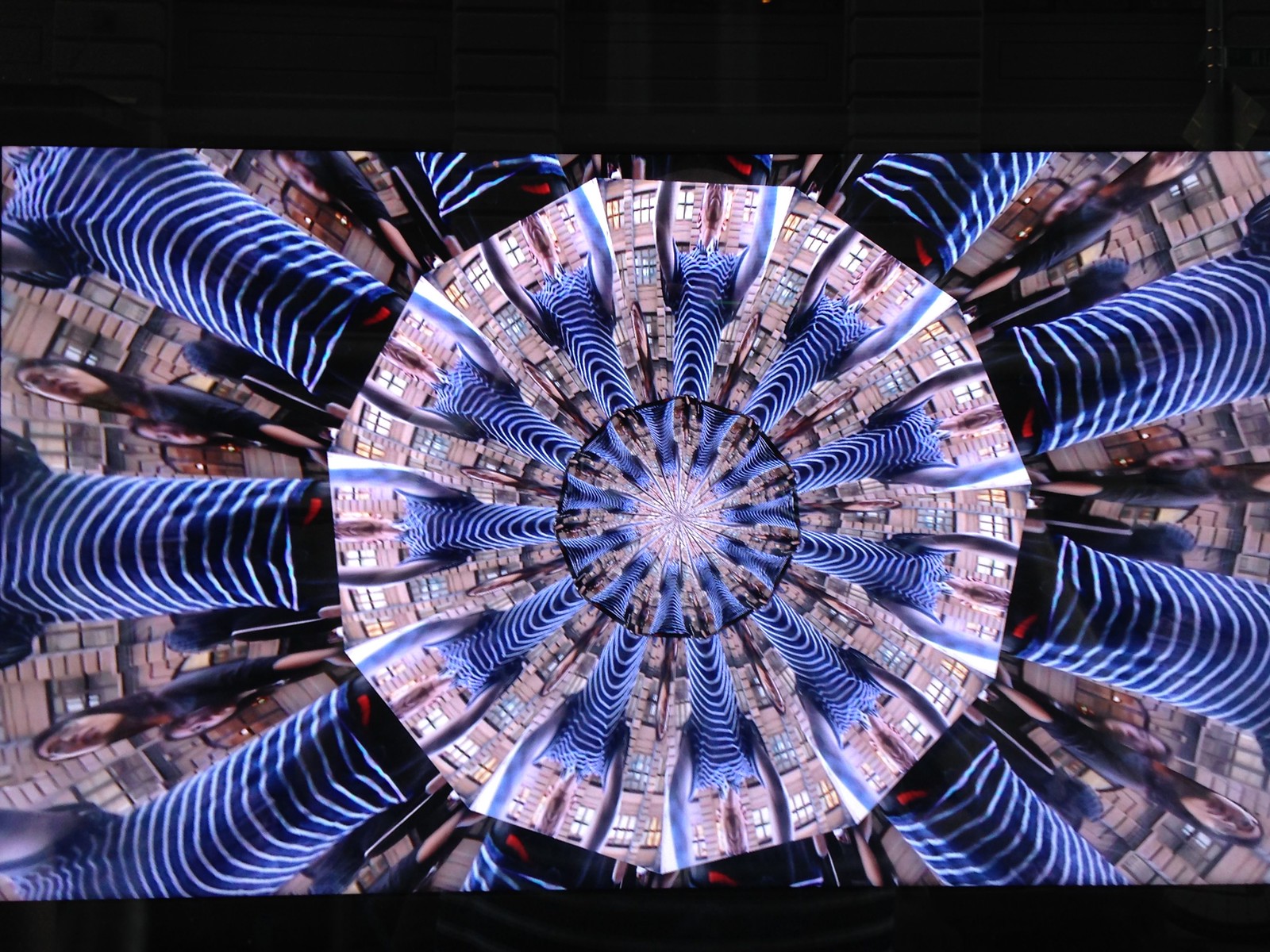 Photograph of a screen showing a kaleidoscope of infinite-mirror-esque concentric discs, each one subdivided into multiple tranches that show the same image, albeit rotated by a consistent amount across the disc. In this case, the image that gets repeated across all discs is a person&rsquo;s black and white striped t-shirt, lending the overall image a very zebra-stripe-like quality.