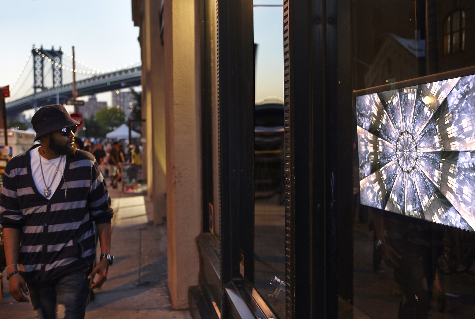 Photograph of a person wearing a dark hat and sunglasses walking up a crowded city block with their head turned towards a storefront window in which a screen displays a kaleidoscopic series of concentric discs, each slice a vibrant, highly contrasted image. Behind the person, far in the background, is a bridge.