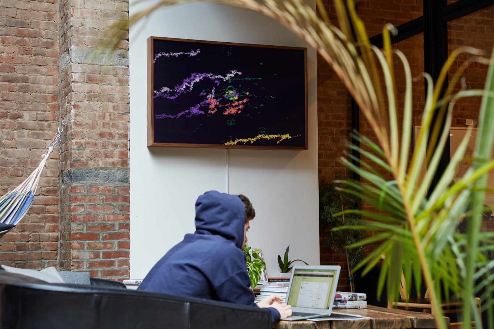 Photograph taken inside a cafe. There's a person with a blue hoodie and a computer at a wooden table in the foreground and an Enter The Machine artwork hanging in the background.
