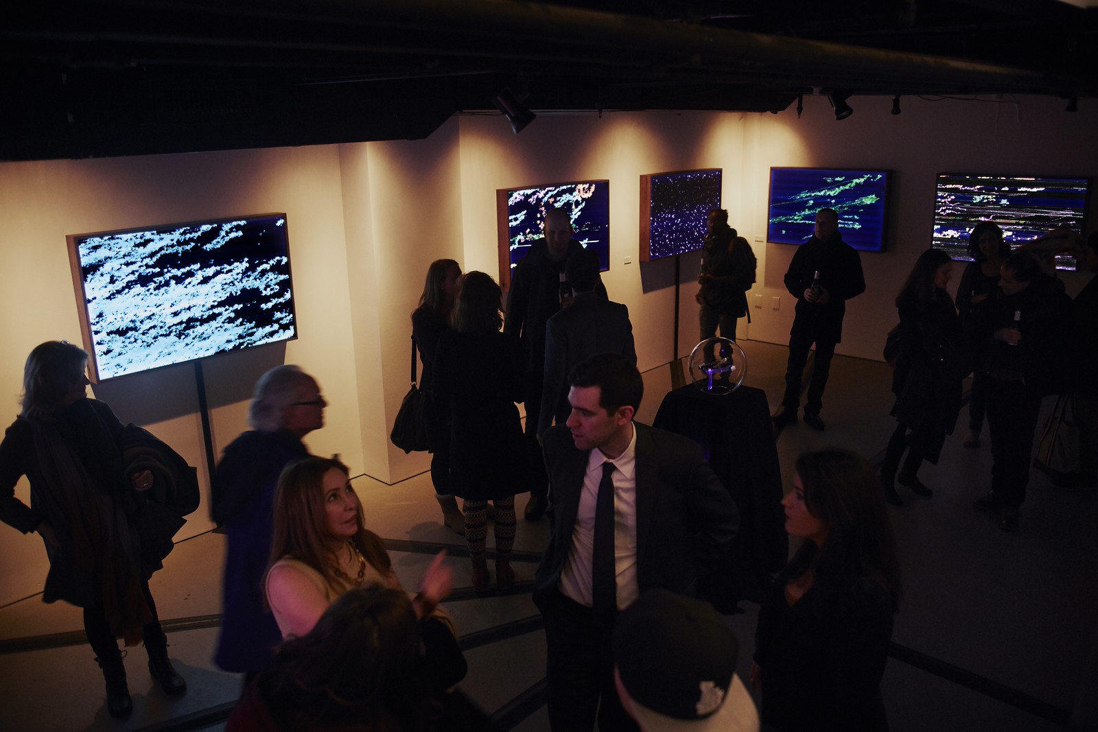 Photograph of a gallery opening taken from above showing roughly 20 people looking at illuminated artworks hanging on a wall; the gallery space is dimly lit.