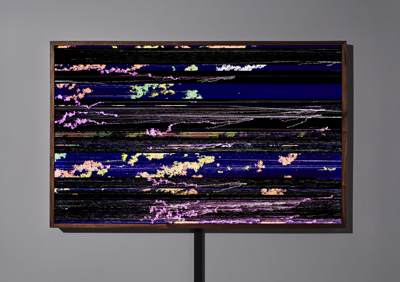 Photograph of a wooden-framed, illuminated artwork hanging on a gray wall showing a glitched-out, strongly horizontal composition of mostly purple and orange organically shaped pixel clusters punctuated by five black-background horizontal rows of varying heights; set against a royal purple background.