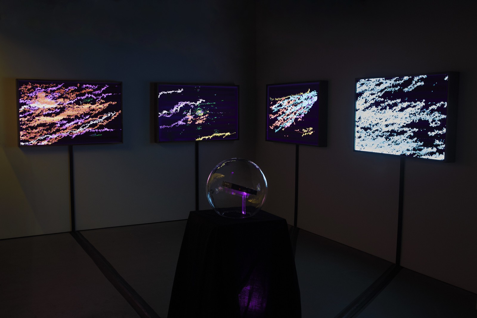 Photograph of a dimly lit, empty gallery space with four illuminated artworks hanging on the wall in the background and a glass globe on a dark pedestal in the foreground.