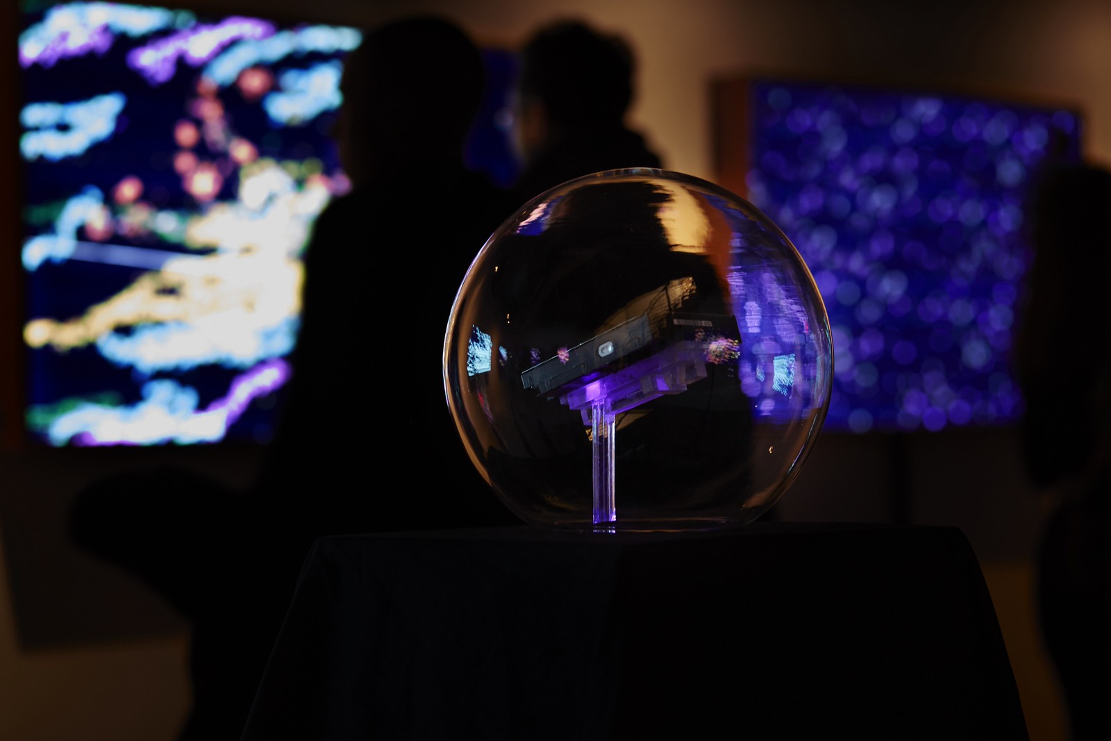 Photograph of a hard drive seemingly floating inside a glass sphere, reflecting the light of the illuminated paintings around it; in the background are two blurry figures looking at artwork.