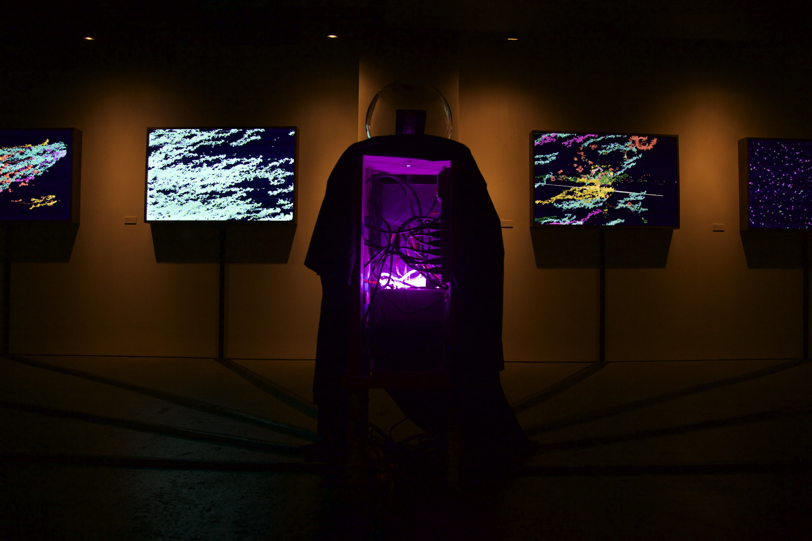 Photograph in a dark art gallery, in the center of which is an open cavity of a box showing electrical wires going hither and thither, backlit by a purple light and shrouded by black cloth on the sides. On top of the box is a glass orb and behind it, flanked on both sides are illuminated artworks hanging on barely lit walls.