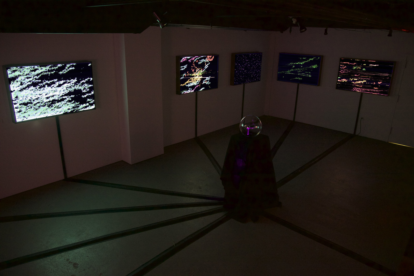 Photograph of a dimly lit, empty gallery space showing five illuminated works hanging on a wall. In the middle of the space is a glass globe on a black pedestal, from the base of which run thick black wire molding that connects to the base of each illuminated artwork.