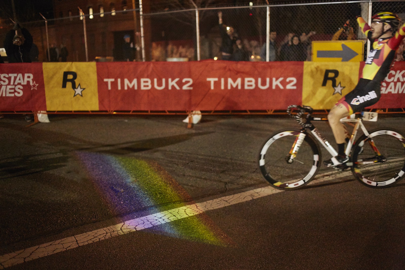 Nighttime photograph of a racing cyclist, hands raised in victory, on the right side of the frame, riding towards a finish line, out of sight on the left side of the frame. In the middle of the race course is a multicolored, striped rectangle that the cyclist is about to ride over.