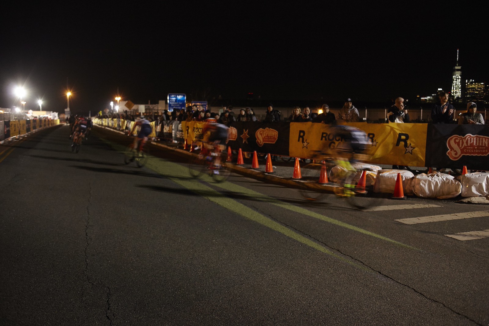 Nighttime photograph of a bike race; several riders are seen rounding a corner and riding over a large, projected yellow arrow.