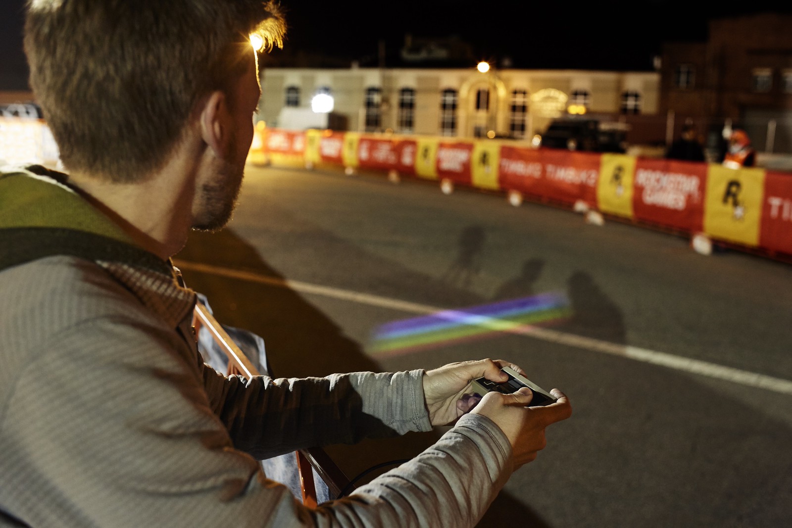 Nighttime photograph of a man, on the left side of the frame, with an (original) Nintendo controller in his hands. On the right side, projected onto the race course behind him, is a multicolored, striped rectangle.