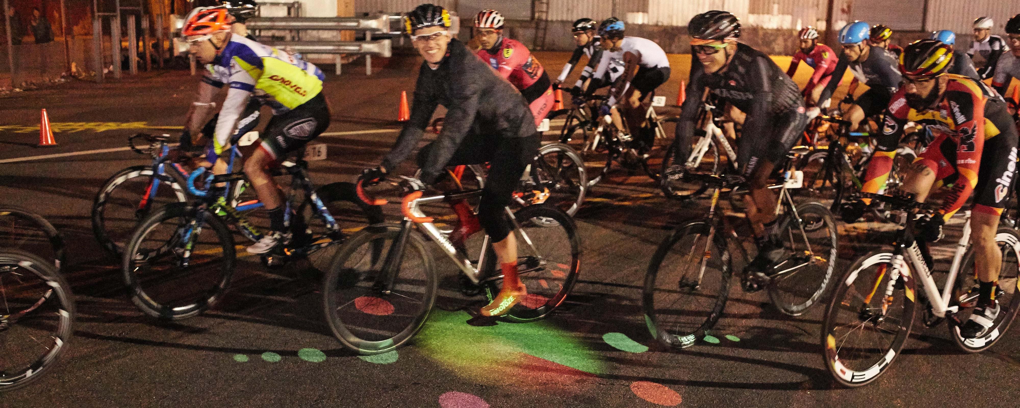Nighttime photograph of a cyclist, in a race, looking straight at the camera (smiling), as he and his bike pass over an illuminated projection of a circular cornucopia of multi-colored shapes that are projected on the race track. He is one of a bunch of cyclists passing over this colorful projection.